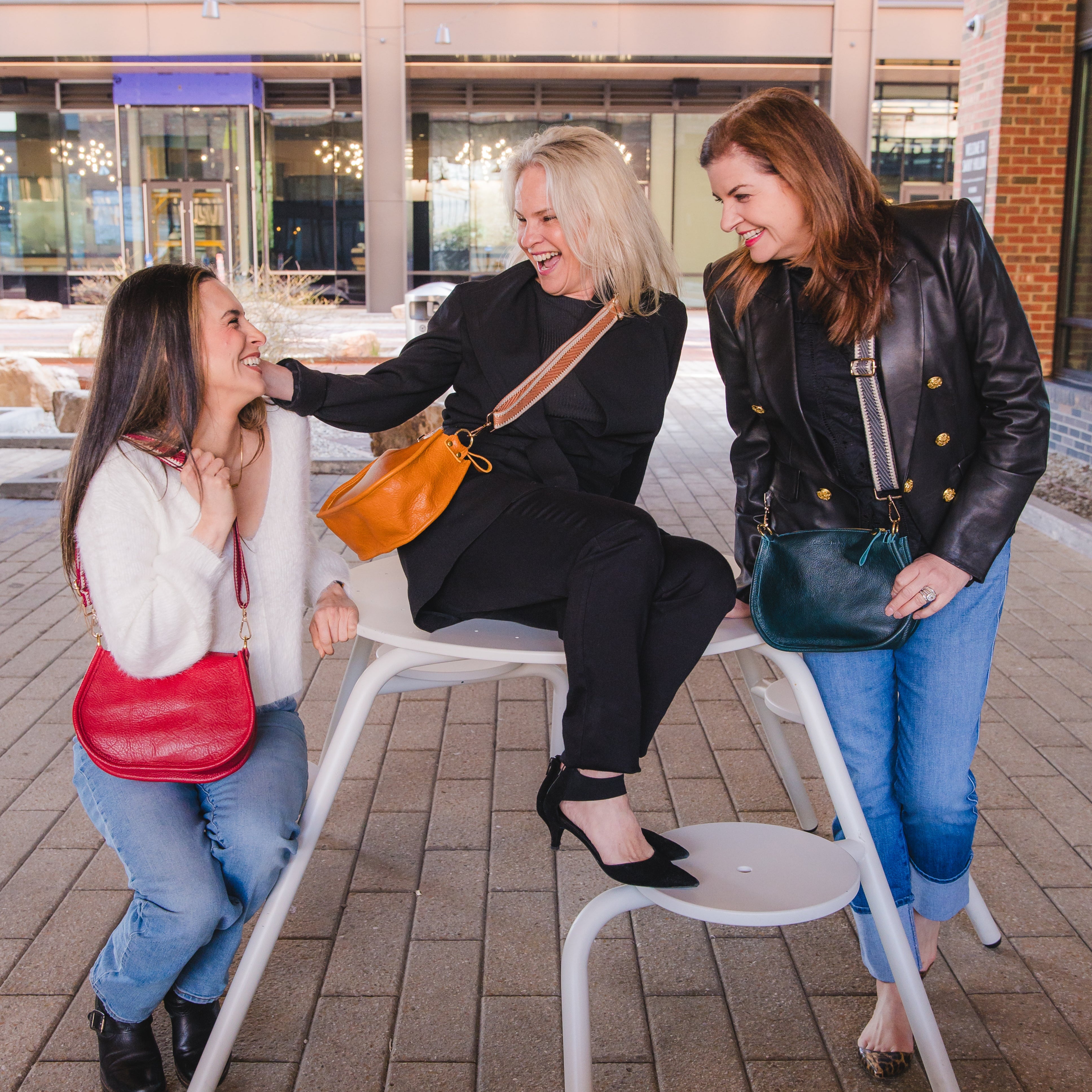 Three women sitting together wearing different color crossbody handbags in the colors red, caramel brown, and teal