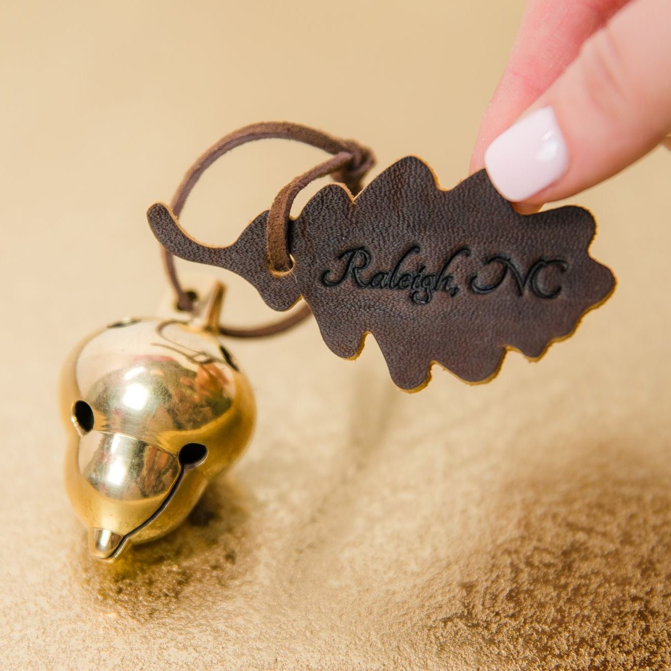 Close-up of a hand holding an acorn-shaped bell ornament with a leather leaf that has 'Raleigh, NC' embossed on it, displayed on a metallic gold surface