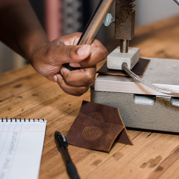 A hand using a tool to emboss a design onto a brown leather piece, with a notebook and pen on a wooden table surface