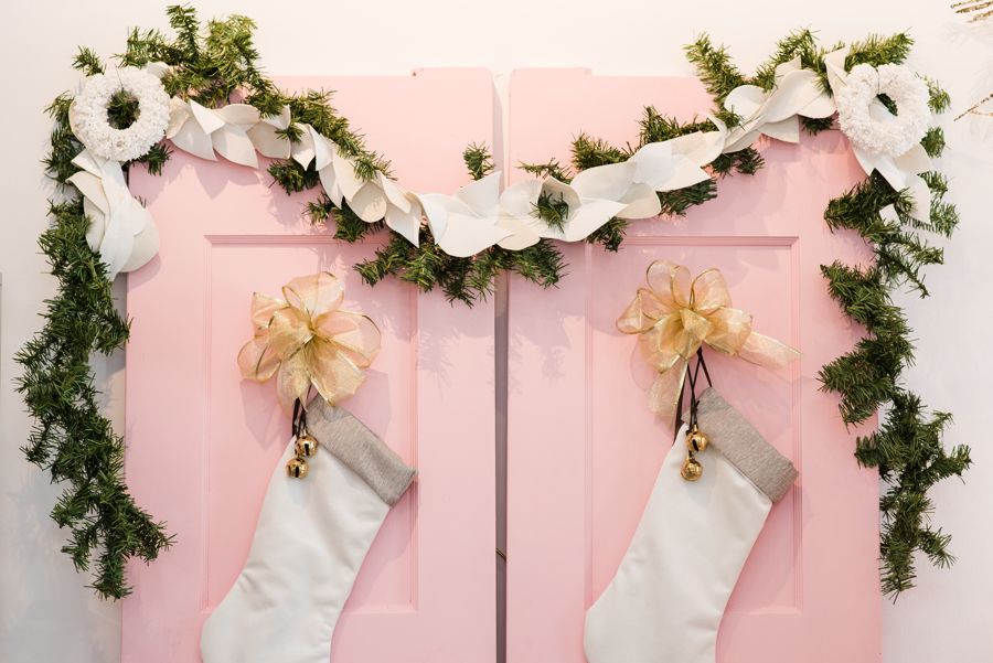 Two jingle bell embellishments adorn white stockings on a pink door with a long wreath hanging above it