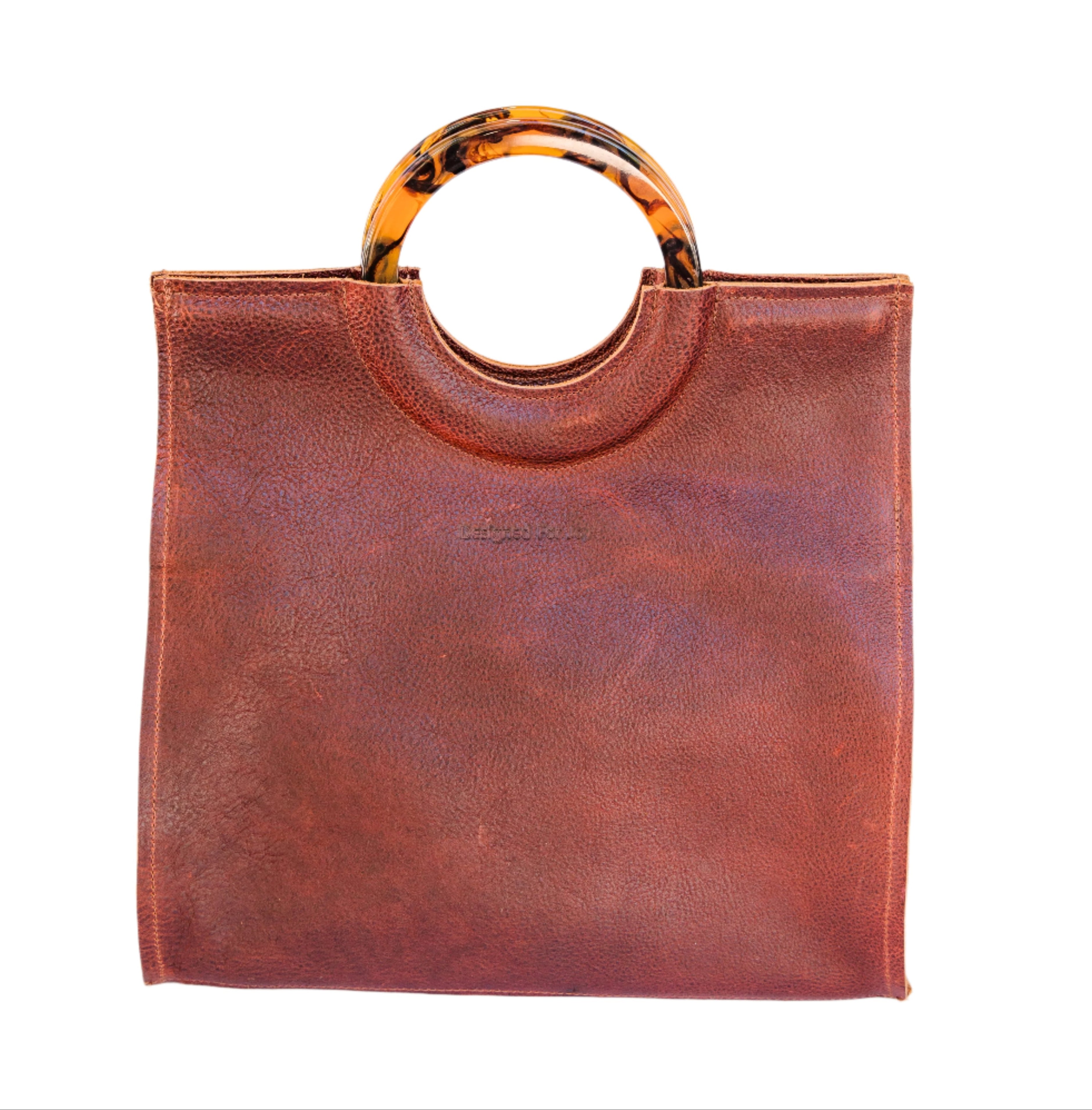 Cinnamon brown leather satchel handbag with round resin handles and a textured surface with 'Designed for Joy' text, displayed on a white background