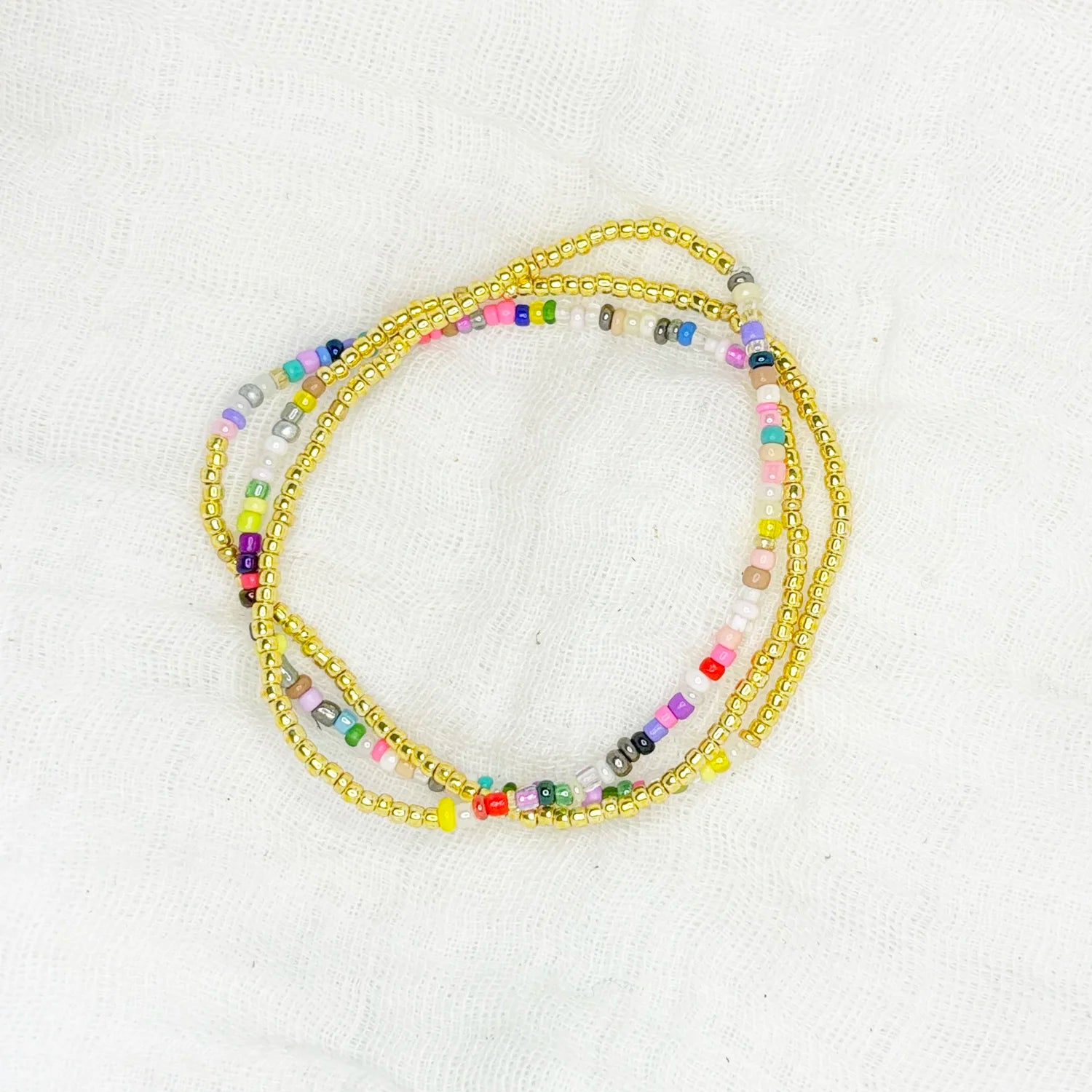 Stackable bracelet with gold seed and multicolored beads, displayed on a white fabric background