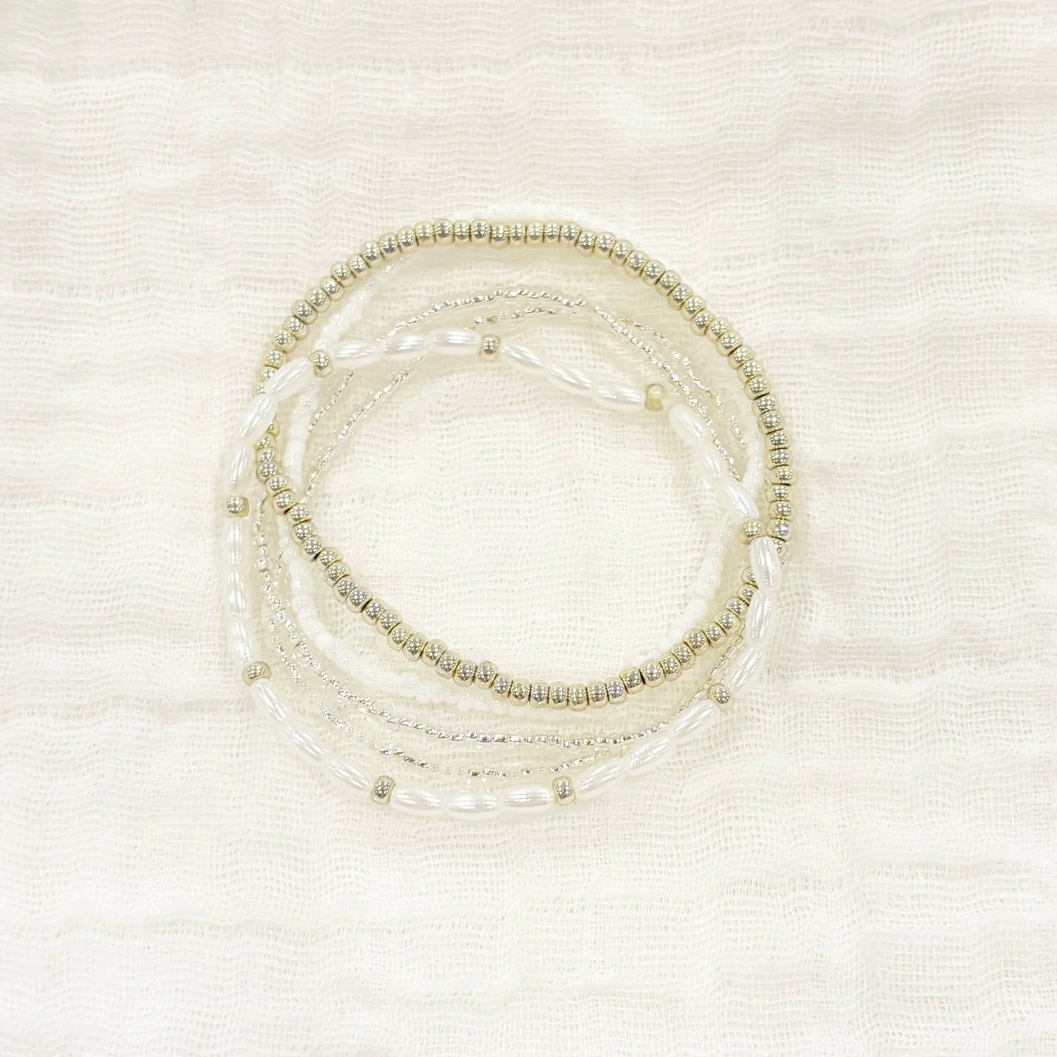 Stackable bracelet with neutral seed, white, and silver beads, displayed on a white fabric background