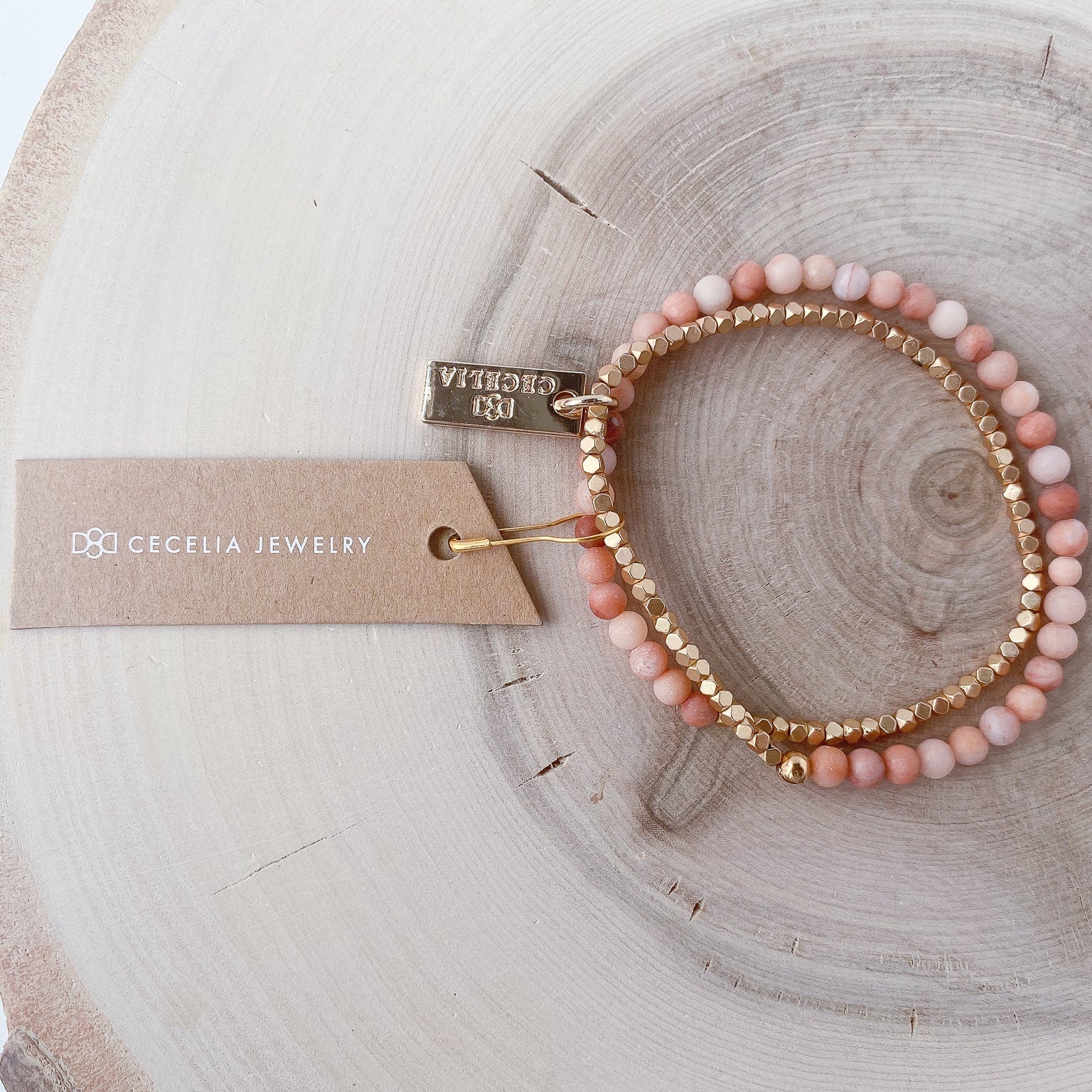 A half peach agate, half matte gold-colored beaded bracelet on a wooden surface with a tag that has 'Cecelia Jewelry' text