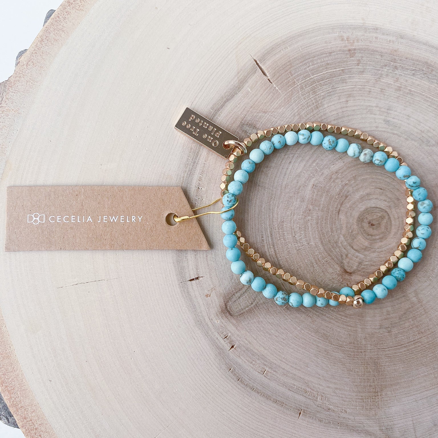 A half turquoise howlite, half matte gold-colored beaded bracelet on a wooden surface with a tag that has 'Cecelia Jewelry' text